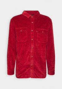 Camisa de pana roja con mangas largas, botón frontal, dos bolsillos en el pecho y un cuello clásico. Tejido texturizado con una sutil canaladura.