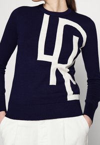 Marinblå tröja i mjukt material med en stor krämfärgad abstrakt logotypdesign. Ribbad krage och fåll med figursydda långärmar.