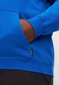 Sweatshirt azul com um bolso frontal, punhos e bainha canelados. Apresenta um pequeno rótulo preto com a inscrição "YOURTURN" perto do bolso. Textura em mistura de algodão.
