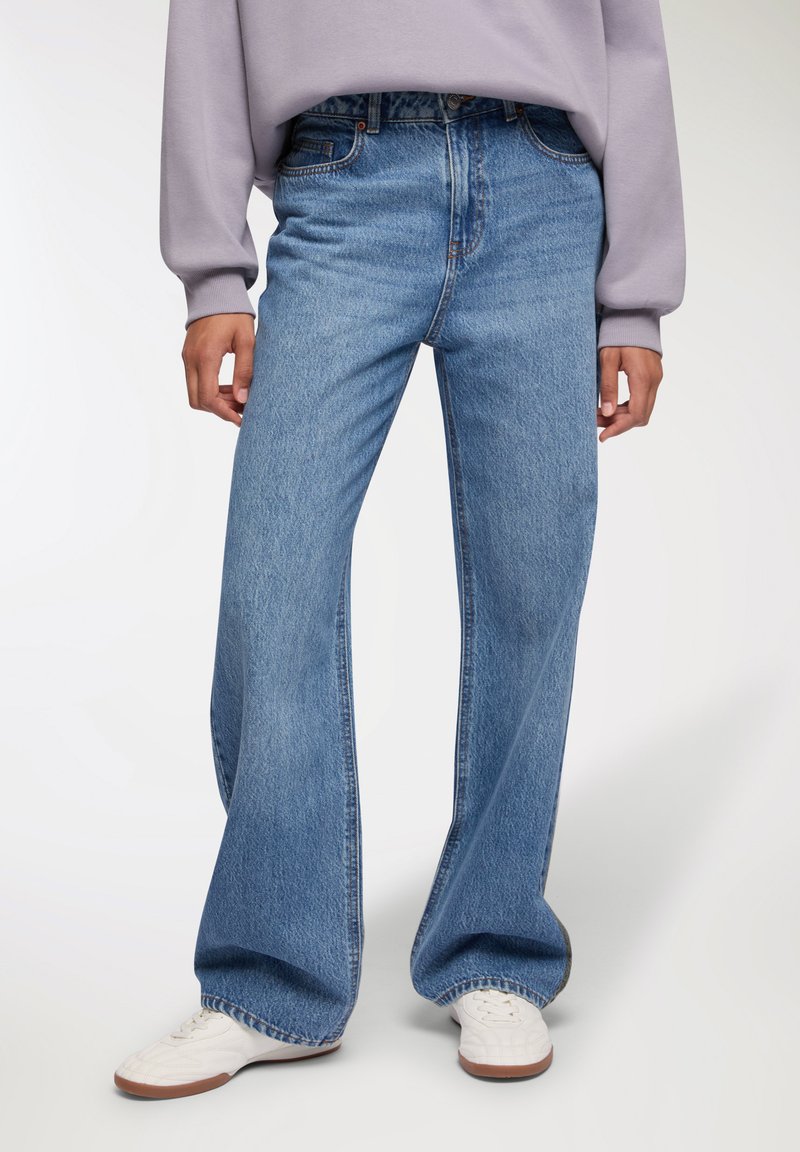 Jeans a gamba larga di un blu chiaro, realizzati in denim, con vita alta, design classico a cinque tasche e leggere sfumature. Indossati con sneakers bianche.