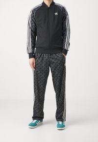 Černá sportovní bunda se světlými pruhy a logem, doplněná kalhotami se vzorem tmavých ptáků. Model má na sobě tyrkysové tenisky.