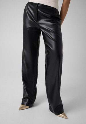 Persona che indossa pantaloni di pelle neri a gamba larga e scarpe con tacco alto dorate e appuntite su uno sfondo neutro.