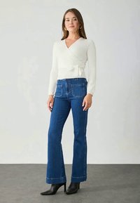 Suéter branco canelado com detalhe de laço, combinado com jeans flare azul-escuro com bolsos frontais e traseiros. O modelo usa botas de tornozelo pretas.