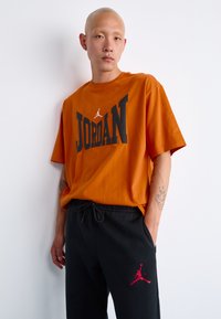Mand med tatoveringer iført en orange Jordan T-shirt og sorte sweatpants med et rødt Jumpman-logo mod en hvid baggrund.