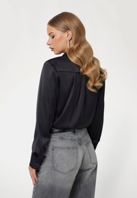 Zwarte satijnen blouse met lange mouwen en een gladde textuur, voorzien van een achterpand en geknoopte manchetten, gecombineerd met vervaagde grijze wijde jeans.