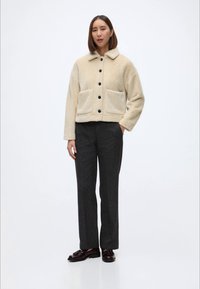 Bežinės vilnos striukės su apykakle, dviem priekinėm kišenėm ir juodais mygtukais, derinama su tamsiai pilkais kelnais ir rudos spalvos bateliais.