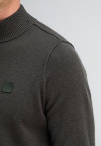 Dunkelgrauer Strickpullover mit hohem Kragen, subtiler Textur und einem kleinen grünen Logopatch auf der linken Seite der Brust.