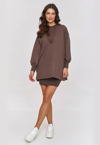 Brązowy, oversize'owy sweter z dekoltem w kształcie litery V, długimi bufkami i teksturowanym wzorem. W zestawie z dopasowaną brązową mini spódnicą i białymi sneakersami.