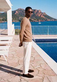 Homme portant des lunettes de soleil et un pull marron se tient sur la terrasse au bord de la piscine avec des chaises longues blanches, de l'eau bleue et des collines rocheuses en arrière-plan.