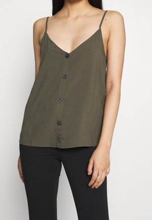 Woman wearing an olive green sleeveless buttoned top with thin straps and black pants, standing against a plain white background.