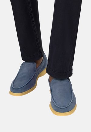 Stopy w niebieskich zamszowych butach wsuwanych z jasnobeżowymi podeszwami, połączone z czarnymi spodniami na jasnym tle.