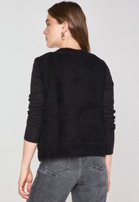 Maglione senza maniche nero con una superficie testurizzata e pelosa e una vestibilità ampia, abbinato a una camicia nera a maniche lunghe e jeans grigi.
