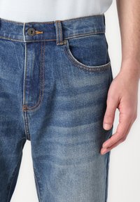 Denim jeans with a faded blue wash, featuring orange stitching, a front button closure, and side pockets. Soft texture and slim fit design.