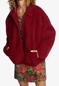 Röd, fluffig fleecjacka med krage, dragkedja och sidofickor, lagd över en färgglad mönstrad klänning. Mjuk textur och oversized passform.