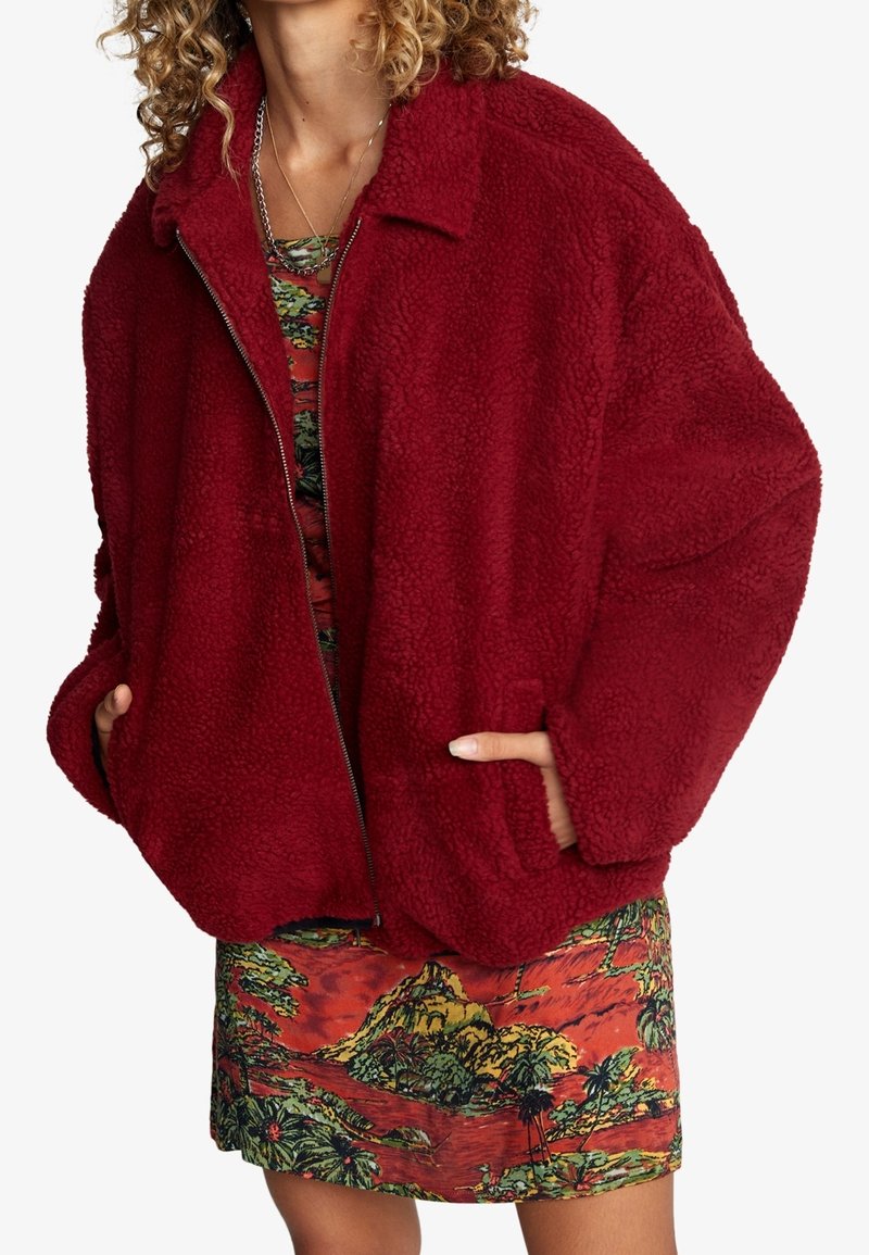 Röd, fluffig fleecjacka med krage, dragkedja och sidofickor, lagd över en färgglad mönstrad klänning. Mjuk textur och oversized passform.