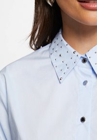 Lichtblauwe button-up shirt met een gespikkelde kraag, versierd met zilveren studs en contrasterende zwarte en blauwe knopen. Glad stoftextuur.