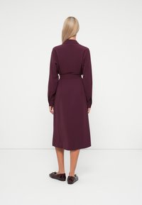 Burgunderfarget midikjole med lange ermer, belte i midjen og glatt tekstur. Kombinert med blanke, mørke sko. Enkel, elegant design.