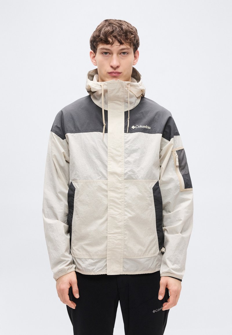 Αντιανεμικό τζάκετ σε μπεζ και μαύρο χρώμα με κουκούλα, εξοπλισμένο με μπροστινό φερμουάρ, ρυθμιζόμενα κορδόνια και τσέπες με φερμουάρ.