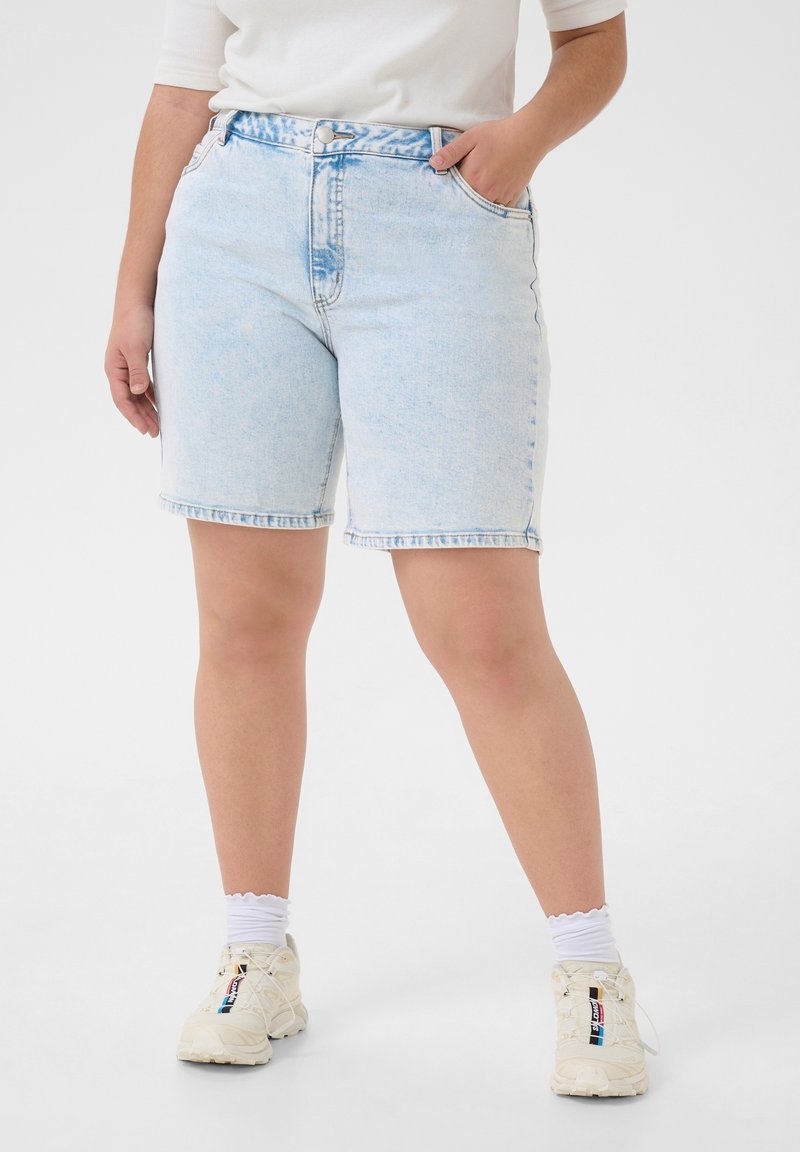 Personne portant un short en jean bleu clair, des chaussettes blanches et des baskets blanches avec une main dans la poche sur un fond uni.