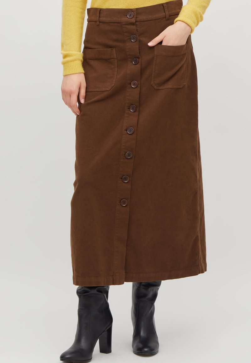 Jupe en denim marron de longueur longue, fermeture à boutons sur le devant et deux grandes poches. Portée avec des bottines noires.