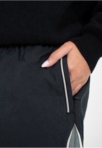 Main avec des ongles rose clair reposant dans la poche d'un pantalon noir avec une bande de finition blanche et grise, portant un haut à manches longues noir.