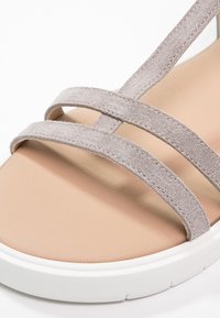 Gros plan d'une sandale beige avec deux brides en daim souple qui se croisent sur une semelle rembourrée et une épaisse semelle blanche.