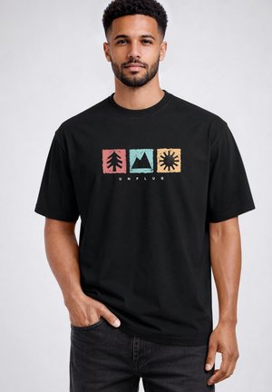 Man with short curly hair wearing a black T-shirt with tree, mountain, and sun symbols in colored squares and the word "UNPLUG" below.