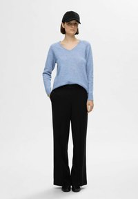 Svetlo modra v-neck pulover iz mehkega blaga, v kombinaciji s črnimi širokimi hlačami in čepico v črni barvi. Model stoji na nevtralni ozadju.