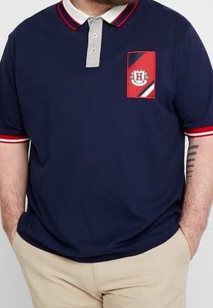 Mand iført navyblå polo med rød- og hvidstribet krave og ærmekanter, beige bukser og en rød emblem på brystet.