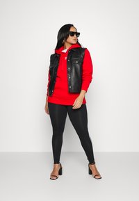 Rood hoodie onder een zwart leren vest, zwarte leggings met witte strepen en zwarte open sandalen met hakken. Zonnebril maakt de look af.