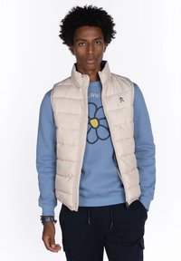 Hombre con cabello rizado que lleva un chaleco de plumón beige sobre una sudadera azul con un gráfico de una gran flor, pantalones oscuros y pulseras en la muñeca izquierda.