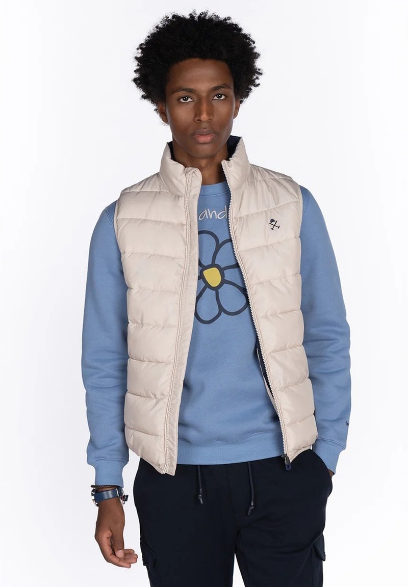 Hombre con cabello rizado que lleva un chaleco de plumón beige sobre una sudadera azul con un gráfico de una gran flor, pantalones oscuros y pulseras en la muñeca izquierda.