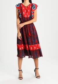 Eine Frau trägt ein rot-blau gemustertes Midi-Kleid mit rüschigen kurzen Ärmeln und schwarzen High Heels mit Knöchelriemen. Sie steht mit einem Arm, der über ihren Körper gekreuzt ist.