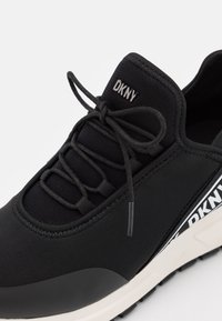 Baskets noires DKNY avec tige en mesh, design à lacets et semelle blanche portant le logo DKNY sur le côté.