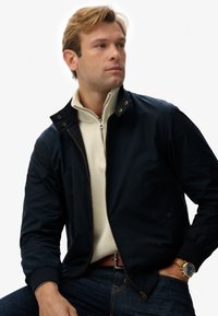 Chaqueta bomber azul marino con cierre frontal, puños y cuello acanalados. Llevada sobre un suéter crema con cremallera, combinada con jeans y un reloj.