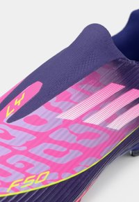 Botas de fútbol con un diseño en púrpura y rosa, textura suave, acentos en rosa llamativo y marca a rayas en el costado.