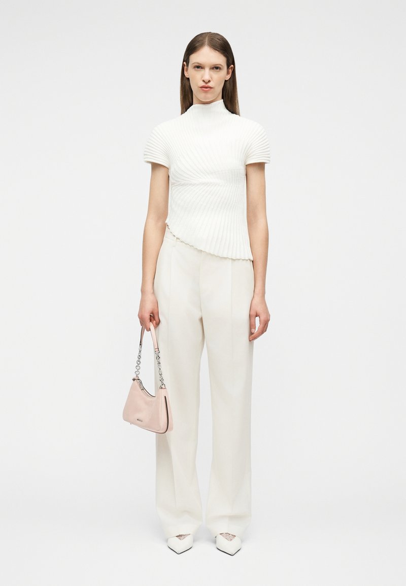 Γυναίκα που φοράει λευκό υφασμάτινο κοντομάνικο μπλουζάκι και παντελόνες με φαρδιά πόδια, κρατώντας μια ανοιχτό ροζ τσάντα χειρός με αλυσίδα, στέκεται μπροστά σε λευκό φόντο.