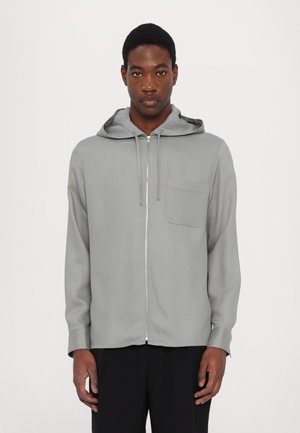 Lys grå zip-up hettegenser i myk stoff med frontlomme og justerbar snorhette, med en jevn tekstur og løs passform.