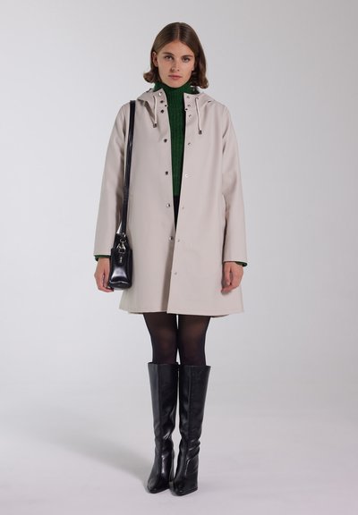 Lichtbeige regenjas met een capuchon en drukknopen aan de voorkant, gedragen over een groene trui. Zwarte kniehoge laarzen en een zwarte schoudertas.