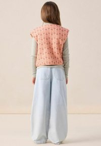 Gilet orange à motifs porté par-dessus une chemise à rayures à manches longues et un Jean large bleu clair, avec des coutures visibles et une coupe décontractée.