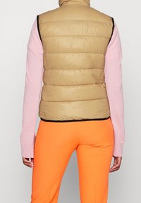 Pletená vesta ve světle hnědé barvě s horizontálním prošíváním, spárovaná s růžovým topem s dlouhými rukávy a oranžovými kalhotami, které mají přiléhavý střih.