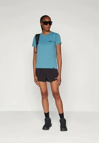 Φωτεινό μπλέ κοντομάνικο μπλουζάκι με λογότυπο, συνδυασμένο με μαύρα αθλητικά σορτς. Το μοντέλο φορά σκούρα γυαλιά ηλίου και μαύρες μποτες πεζοπορίας.