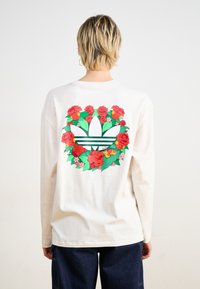 Dugih rukava krem majica s cvjetnim uzorkom crvenih ruža oko središnjeg logotipa u zelenoj i bijeloj boji, uz dodatne šarene akcenti.