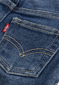 Kieszeń dżinsowa Levi's z klasycznym niebieskim materiałem, żółtym przeszyciem i czerwoną etykietą. Kieszeń ma charakterystyczny, zakrzywiony kształt.