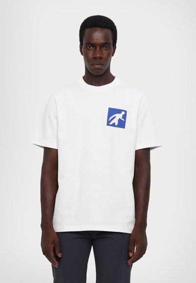Vit t-shirt i bomull med en blå fyrkantig grafisk design som har en vit figur på vänster bröstområde. Kort ärm och rund halsringning.