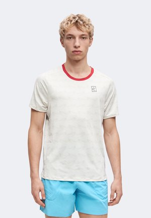 Ung mand med krøllet blondt hår iført hvidt mønstret t-shirt med rød krave og lyseblå shorts, vendt mod kameraet foran ensfarvet baggrund.