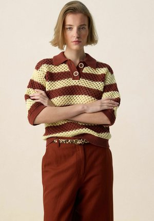 Jonge vrouw met kort haar die een bruin- en crèmekleurig gestreept gebreid bovenstuk met knopen draagt, roestkleurige broek en een luipaardprint riem, met gekruiste armen.