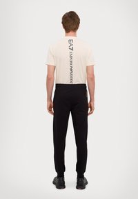 Világos bézs színű póló függőleges fekete logóval, slim fit fekete jogger nadrággal párosítva, állítható derékkal és bordázott mandzsettákkal.