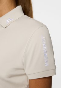 Jasno beżowa koszulka polo wykonana z gładkiego materiału. Posiada ściągaczowy kołnierz oraz haftowane logo 'J.LINDEBERG' na rękawie.
