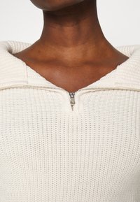 Cremfarbener Strickpulli mit strukturiertem Muster, der über einen halben Reißverschluss und eine Kapuze verfügt. Der Stoff wirkt weich und gemütlich.